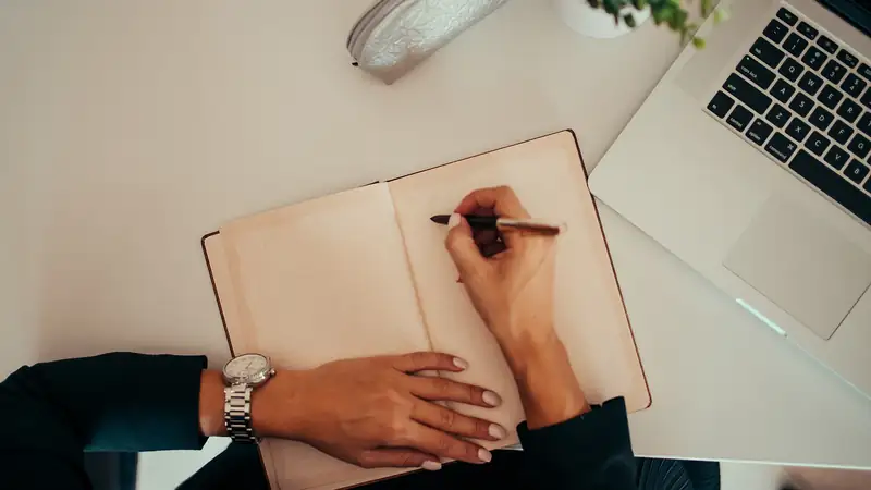 Person, die neben einem Laptop in ein Notizbuch schreibt